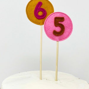May include: Two felt cake toppers, one with the number 6 in pink on a gold circle and the other with the number 5 in brown on a pink circle.