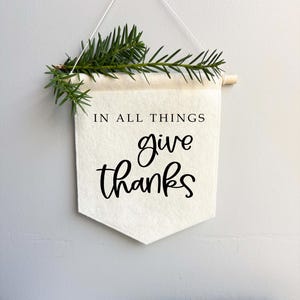 May include: A white felt banner with the words "IN ALL THINGS give thanks" in black script. A small wooden dowel and a sprig of green pine needles are attached to the top, creating a rustic, minimalist wall hanging.