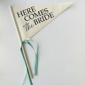 May include: A white fabric pennant with a wooden stick and teal ribbons. The pennant has black text that reads "Here Comes the Bride."