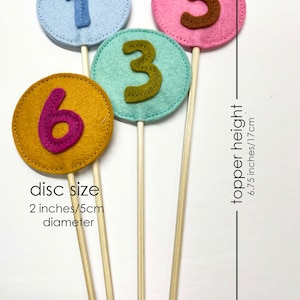 May include: Four felt cake toppers with numbers 1, 3, 5, and 6. The numbers are in different colors and are attached to wooden sticks. The toppers are 6.75 inches tall and the discs are 2 inches in diameter.