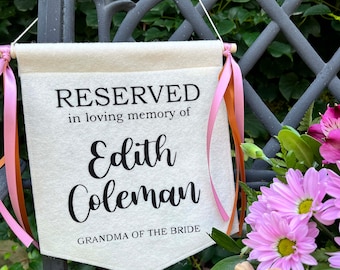 May include: A white fabric banner with pink and orange ribbons hanging from it. The banner reads "RESERVED in loving memory of Edith Coleman GRANDMA OF THE BRIDE".