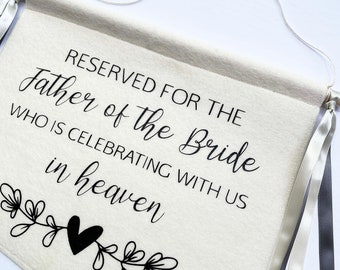 May include: A white felt banner with black text that reads "Reserved for the father of the bride who is celebrating with us in heaven". The banner has two black and two silver ribbons attached to the top.