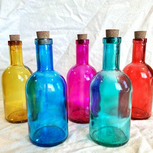 Puede incluir: Cinco botellas de vidrio de colores con tapones de corcho. Las botellas son en tonos de amarillo, azul, rosa, verde azulado y rojo. Las botellas están dispuestas sobre una superficie blanca.