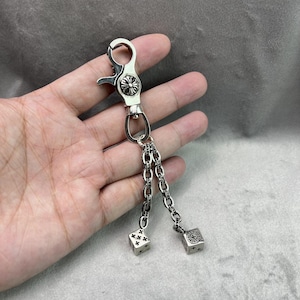 May include: Silver-toned keychain with a clasp, decorative flower, and two chains each ending in a die. The dice have black markings. The keychain is held in a hand.