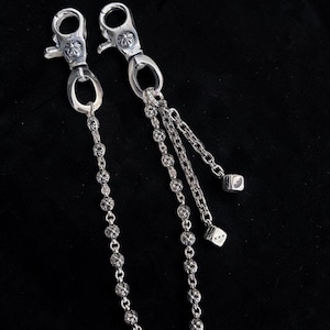 Op de afbeelding: Twee zilverkleurige metalen portemonnee kettingen met karabijnsluitingen. De ene ketting heeft decoratieve kralen, de andere heeft drie strengen en twee dobbelsteen bedels. De kettingen worden tegen een donkere achtergrond getoond.