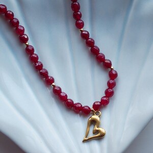 May include: A red beaded necklace with a gold heart pendant. The necklace has small, faceted red beads and gold accents. The heart pendant is a simple, open design. The necklace is displayed on a white, scalloped dish.