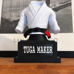 Pode incluir: Uma parte superior de gi de artes marciais branca com uma faixa preta com ponta vermelha, exibida em um suporte preto. A base do suporte é retangular e apresenta uma placa preta com o texto branco TUGA MAKER centralizado na frente.