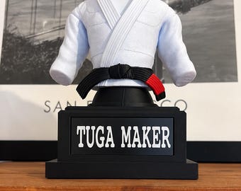 May include: A white martial arts gi top with a black belt featuring a red tip, displayed on a black stand. The base of the stand is rectangular and features a black plaque with the white text TUGA MAKER centered on the front of the display.