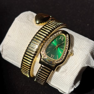 May include: A gold-toned wristwatch with a green face and a diamond-studded bezel. The watch has a snake-like design, with a flexible band that wraps around the wrist. The watch is resting on a white surface.