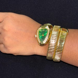 May include: A gold-toned wristwatch with a green face and diamond accents, styled in a snake design. The watch is paired with a matching gold bracelet, both worn on a wrist. The background is a dark, solid color.