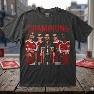 May include: Dark gray t-shirt with a graphic of a team holding a trophy, the word "CHAMPIONS" in red, and the text "GUNNERS PRIDE WEAR". The team is wearing red and white jerseys with the Emirates logo.