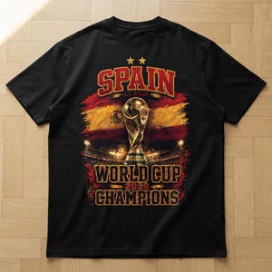 Può includere: Una t-shirt nera con una stampa grafica del trofeo della Coppa del Mondo centrata su uno sfondo con la bandiera spagnola. Il testo recita SPAIN, LA ROJA, WORLD CUP 2026 e CHAMPIONS in caratteri audaci e stilizzati con due stelle dorate.