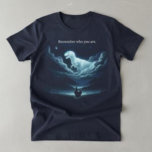 Puede incluir: Camiseta azul marino con un diseño surrealista. Muestra un dinosaurio emergiendo de las nubes sobre una gallina, con el texto "Remember who you are." El diseño incluye una luna y un paisaje.