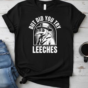 Puede incluir: Camiseta negra con un gráfico blanco de un médico de la peste y el texto "BUT DID YOU TRY LEECHES". El diseño muestra un médico de la peste con sombrero y máscara. La camiseta está doblada en la parte inferior.