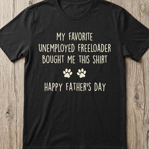 Könnte beinhalten: Schwarzes T-Shirt mit weißem Text: "MY FAVORITE UNEMPLOYED FREELOADER BOUGHT ME THIS SHIRT" und "HAPPY FATHER'S DAY" mit Pfotenabdrücken. Ein lässiges, bequemes T-Shirt für Vatertag.