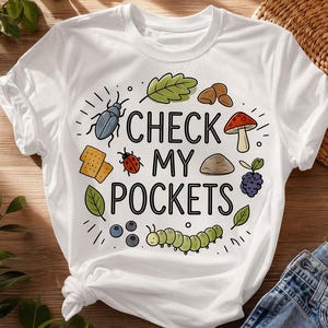Peut inclure: T-shirt blanc avec le texte "CHECK MY POCKETS" et des illustrations d'un coléoptère, d'une coccinelle, d'une feuille, d'un champignon, de noix, d'une pierre, de baies, de myrtilles, de crackers et d'une chenille. Le t-shirt est en matière souple.
