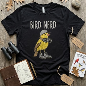 May include: Black t-shirt with the embroidered text "BIRD NERD" above a cartoon yellow bird wearing a cap, binoculars, and boots. Accessories include a notebook, pen, binoculars, and a bird guide.