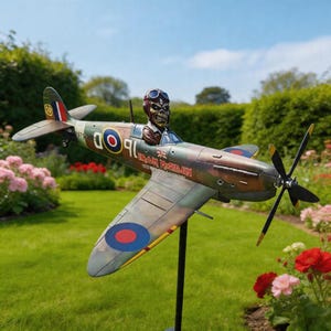 Può includere: Un modello di aereo Spitfire con elica e la scritta "Iron Maiden" sul lato. L'aereo presenta un motivo mimetico con dettagli rossi, blu e gialli. Una figura con casco e occhiali da pilota è nell'abitacolo. L'aereo è montato su un supporto nero.