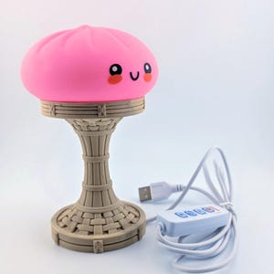 Peut inclure: Une lampe rose arrondie en forme de ravioli avec un visage souriant, dotée d'yeux noirs et d'une petite bouche. La lampe repose sur une base beige texturée rappelant un panier tressé, reliée à un câble d'alimentation USB blanc avec télécommande.