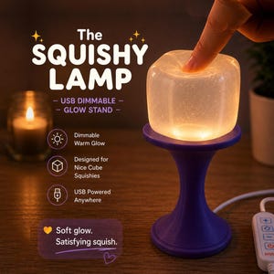 May include: A squishy, translucent, cube-shaped lamp emitting a warm glow, resting on a purple pedestal stand. The lamp is USB-powered with an inline controller. Text on the image reads: The SQUISHY LAMP, USB DIMMABLE GLOW STAND, Dimmable Warm Glow, Designed for Nice Cube Squishies, USB Powered Anywhere, Soft glow. Satisfying squish.