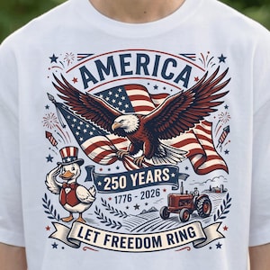 以下が含まれることがあります： アメリカの250周年を祝う愛国的なデザインの白いTシャツ。グラフィックには、ワシ、アメリカ国旗、愛国的な衣装を着たアヒル、トラクター、そして「AMERICA」、「250 YEARS 1776-2026」、「LET FREEDOM RING」の文字が含まれています。
