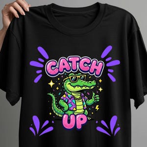 May include: A black t-shirt featuring a graphic of a green crocodile wearing yellow sunglasses, a gold chain, and a patterned shirt. The crocodile is giving a thumbs up. The text CATCH UP is printed in large pink letters above and below the crocodile.