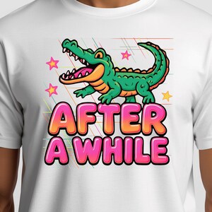 May include: A white t-shirt featuring a graphic of a green crocodile with an orange underbelly, surrounded by pink stars and thin diagonal lines. Below the crocodile, the text "AFTER A WHILE" is printed in large, bubble-style pink and orange letters.
