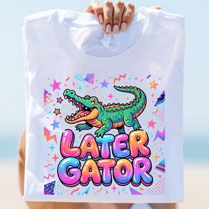 May include: A white t-shirt featuring a colorful graphic of a green alligator with an open mouth. Below the alligator, the text LATER GATOR is printed in large, bubble-style letters with a gradient of pink, orange, and blue. The design includes stars.