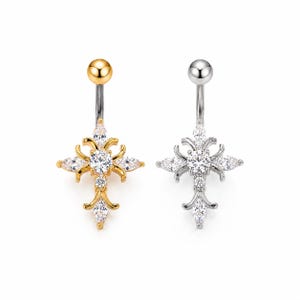 Könnte beinhalten: Zwei Bauchnabelpiercings, eines goldfarben und eines silberfarben, jeweils mit einem kreuzförmigen Design. Die Kreuze sind mit klaren, funkelnden Edelsteinen verziert. Der goldene Ring hat eine goldene Kugelspitze, der silberne Ring hat eine silberne Kugelspitze.