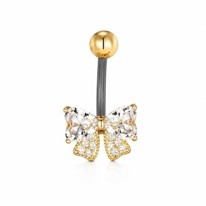 May include: A gold-toned belly button ring featuring a bow design. The bow is adorned with clear, sparkling crystals and smaller accent stones. The top ball is gold, and the barbell is dark gray.