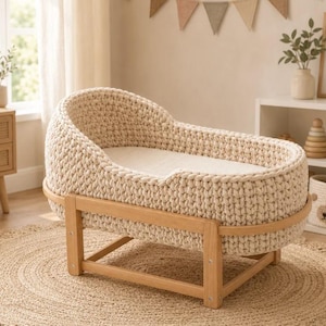 May include: A cream-colored, chunky knit baby bassinet featuring a thick, textured woven exterior. The oval-shaped basket rests securely on a light-toned wooden frame with four sturdy legs and a smooth, flat base for support.