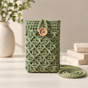 Puede incluir: Una funda de teléfono de ganchillo verde salvia con un patrón texturizado y un cierre de botón de madera. La funda tiene una correa a juego y está diseñada para guardar un teléfono inteligente. Un accesorio elegante.