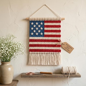May include: A handmade macrame wall hanging depicting the American flag. The flag features a blue field with white stars, red and white stripes, and a fringe at the bottom. A tag reads "Beginner PDF Pattern".