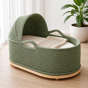 May include: A sage green crocheted bassinet with a rounded hood and integrated handles. The interior features a smooth off-white mattress pad topped with a folded beige knit blanket. The structure rests on a light-toned wooden oval base.