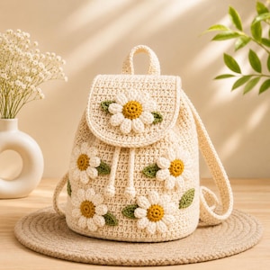 May include: Cream-colored crochet backpack adorned with daisy flower appliqués. The daisies have white petals, yellow centers, and green leaves. The bag features a drawstring closure, a flap, and adjustable straps. The backpack is sitting on a round, woven mat.