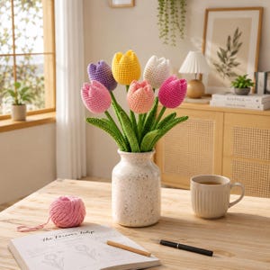 Könnte beinhalten: Ein Strauß handgehäkelter Tulpen in einer gesprenkelten Vase. Die Tulpen sind in verschiedenen Farben gehalten: rosa, lila, gelb, pfirsich, weiß und fuchsia. Die Vase steht auf einem Holztisch mit einem Knäuel rosa Garn, einem Buch und einer Tasse Tee.