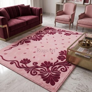 May include: A rectangular area rug featuring a light pink base with a dark burgundy floral scroll pattern along the edges and scattered petal motifs across the center. The text RugsArt is printed in the middle of the rug surface.