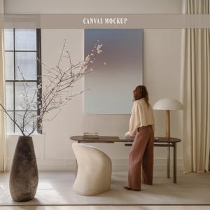 May include: A room interior featuring a canvas mockup with the text "CANVAS MOCKUP". The room includes a large window, a dark wooden desk, a white chair, and a tall vase with branches. A person is standing near the desk.