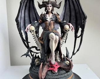 Puede incluir: Figura detallada de una demonio femenina sentada en un trono. La figura tiene grandes alas negras, cuernos y un vestido oscuro con detalles dorados. El trono está adornado con calaveras y una base roja y puntiaguda, todo sobre una plataforma circular.