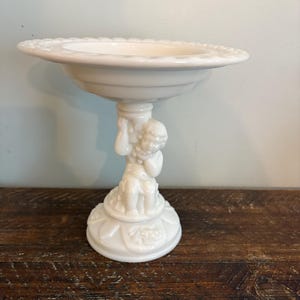 Puede incluir: Una fuente blanca para servir sobre pedestal. El plato tiene un borde ancho y festoneado y está sostenido por una figura de querubín. La base es redonda con un patrón decorativo. El plato está hecho de un material brillante y opaco.