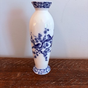 Könnte beinhalten: Weiße Keramikvase mit blauem Blumen- und Vogelmuster. Die Vase hat eine schlanke, längliche Form mit ausgestelltem Ober- und Unterteil. Das blaue Design umfasst Zweige, Blumen und Vögel, die eine klassische, elegante Ästhetik schaffen.