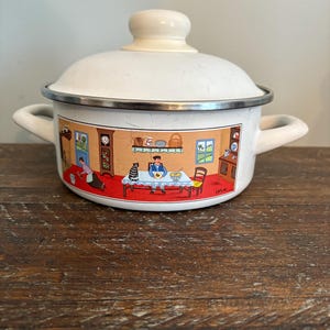 Puede incluir: Olla de esmalte blanco con tapa y dos asas. La olla presenta una colorida ilustración de una escena de cocina alrededor de la base. La tapa y las asas son de color crema, y el borde es plateado. La olla está sobre una superficie de madera.