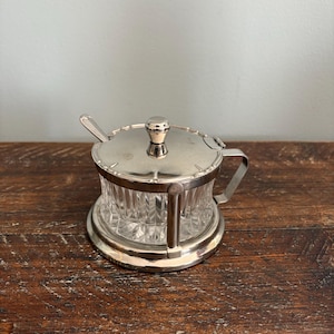 Op de afbeelding: Een verzilverde suikerpot met een doorzichtige, facetgeslepen glazen container. De pot heeft een deksel met een kleine handgreep, een handgreep aan de zijkant en een kleine lepel. De pot staat op een ronde basis.