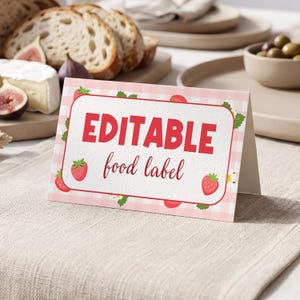 May include: A folded, rectangular food label with a pink gingham pattern and a red border. The label features the words "EDITABLE food label" in red and a strawberry illustration. The label is set on a beige linen tablecloth.