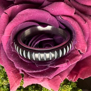 Könnte beinhalten: Ein silberfarbener Ring mit schwarzem Band und weißen Akzenten, eingebettet in eine tiefviolette Rose. Das Ringdesign zeigt vertikale weiße Linien und rechteckige Formen. Die florale Anordnung sorgt für einen auffälligen Kontrast.