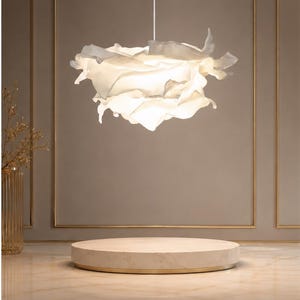 Peut inclure: Un luminaire suspendu blanc en forme de fleur est suspendu au-dessus d'une plateforme d'exposition ronde et beige. La lumière a un design en couches, en forme de pétales. L'arrière-plan présente un mur neutre avec une bordure dorée.