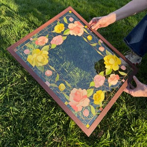 May include: A square mirror with a dark brown wooden frame. The mirror's surface is decorated with a floral design featuring yellow and pink flowers, green leaves, and gold accents. The mirror is held by two hands.
