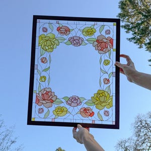 May include: A rectangular stained glass panel with a dark brown frame, held against a blue sky. The panel features a floral design with yellow, pink, and red roses, and green leaves. The glass has a clear center.