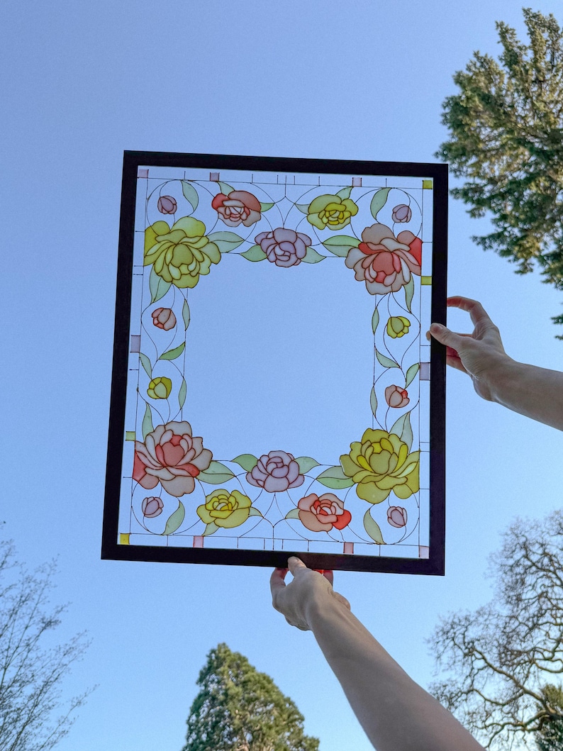 May include: A rectangular stained glass panel with a dark brown frame, held against a blue sky. The panel features a floral design with yellow, pink, and red roses, and green leaves. The glass has a clear center.
