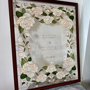May include: A framed artwork featuring white floral designs and the names "EMILY & DIMITRI" with the date "09.05.26". The frame is a dark brown color, and the artwork has a transparent background.